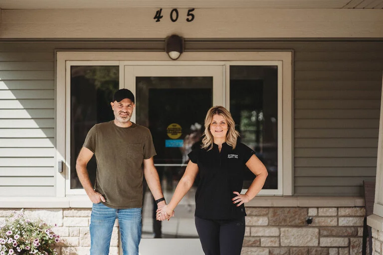 A man and woman stand together outside a house, smiling and enjoying a sunny day in a friendly neighborhood setting.