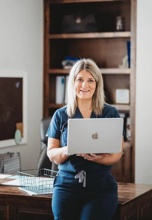 A woman in medical scrubs is holding a laptop, appearing focused and engaged in her work environment.